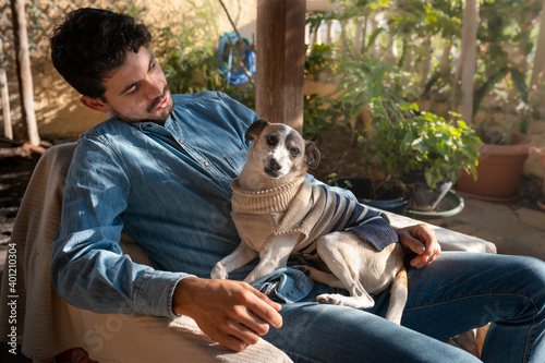 young man sitting on a chair in a garden interacts with gray dog ​​sitting on him.