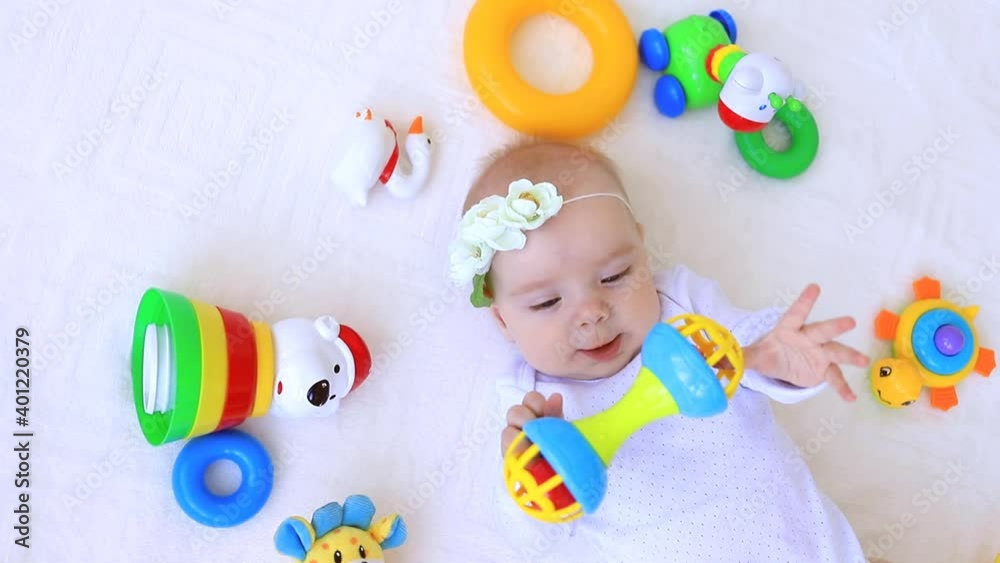 little baby playing with rattles on bed at home in white clothes among toys
