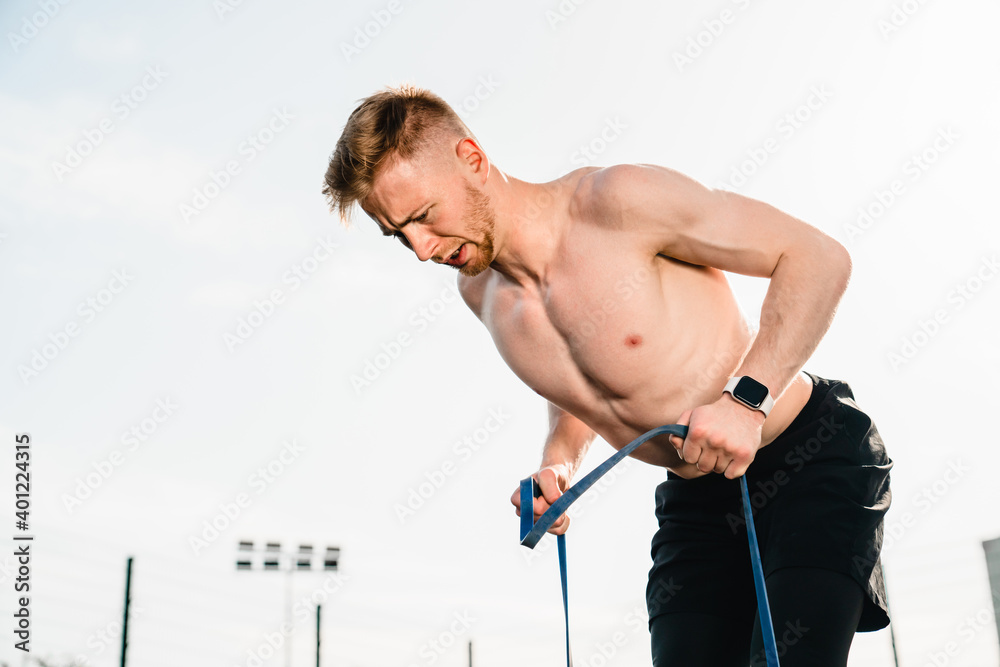 Sporty young shirtless athlete training with rubber band outdoors