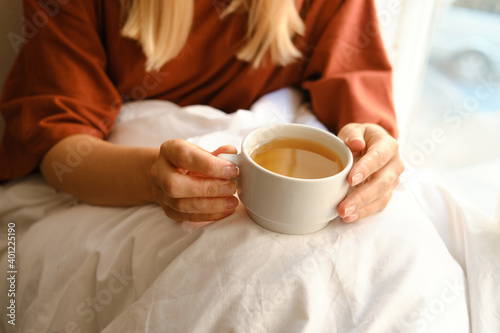 The girl drinks tea in the morning near the window. A cup of hot tea in hands. Awakening from sleep. The happy woman is wrapped in a blanket. Dreams and think about life. Enjoy cozy in home