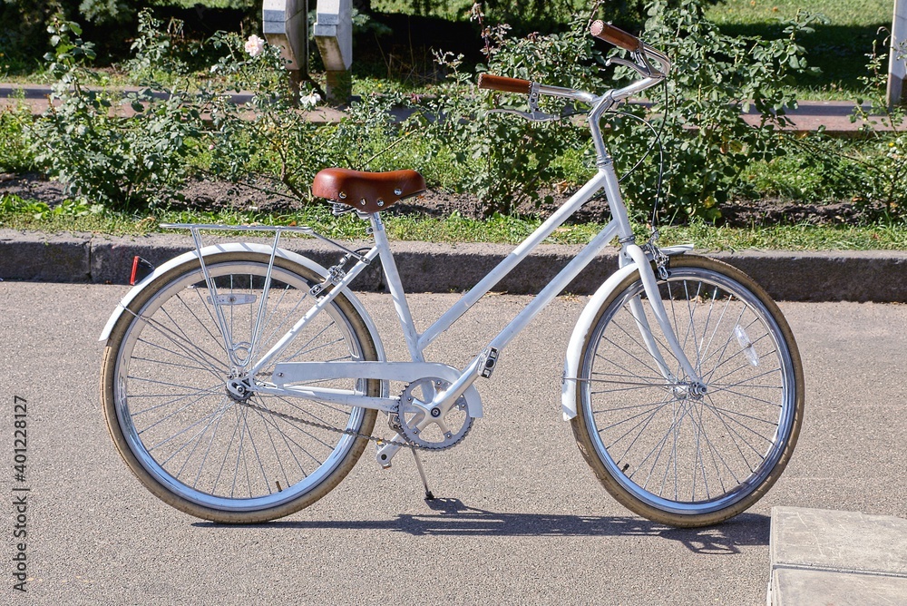 one old white female bicycle with a brown leather saddle stands on a gray asphalt road in the park against a background of green vegetation