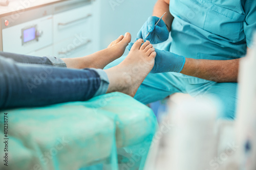 Podiatry specialist in blue gloves cleaning skin of client from callus and corns with professional tools