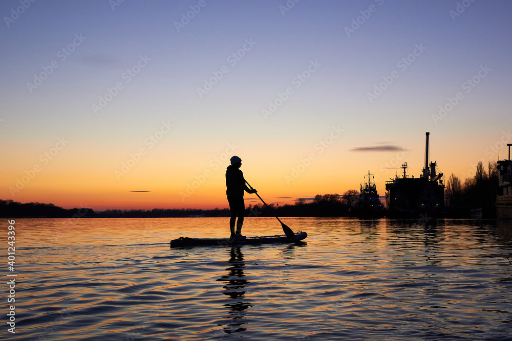 A middle-aged woman on stand up paddle boarding at dusk on a flat quiet winter river with beautiful sunset colors
