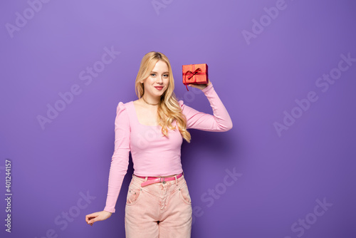 happy blonde young woman holding red gift box on purple background.