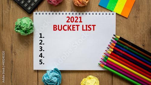 2021 bucket list symbol. White note with inscription '2021 bucket list' on beautiful wooden table, colored paper, colored pencils, clips, coins and calculator. Business and 2021 bucket list concept. © Dzmitry