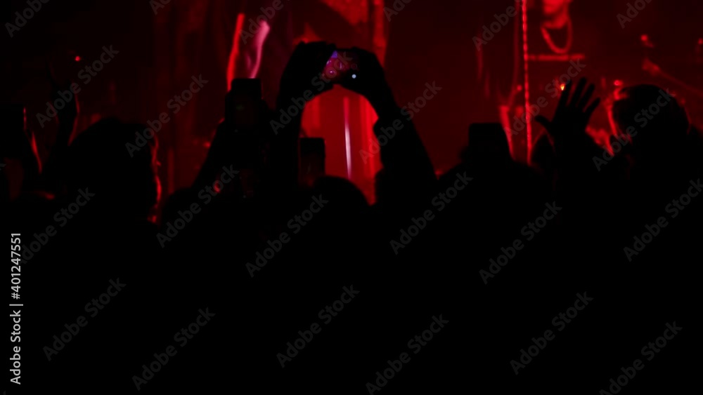 People in front of the scene watch music concert and dance in slow motion. Close-up of spectators who jump with hands up and listen to a popular band in a night club. They are making video on mobiles.