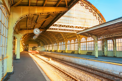 View of the old railway station with a large metal arch and an empty platform with a clock for people on a clear sunny day.