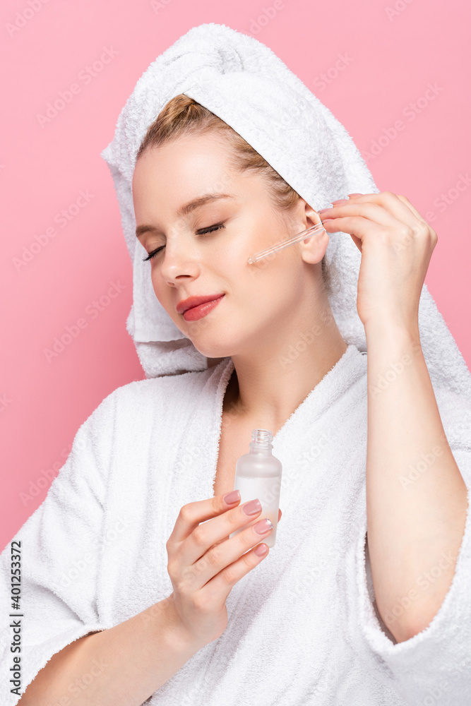 young woman in bathrobe with towel on head applying serum on face isolated on pink. young woman in bathrobe with towel on head applying serum on face isolated on pink.