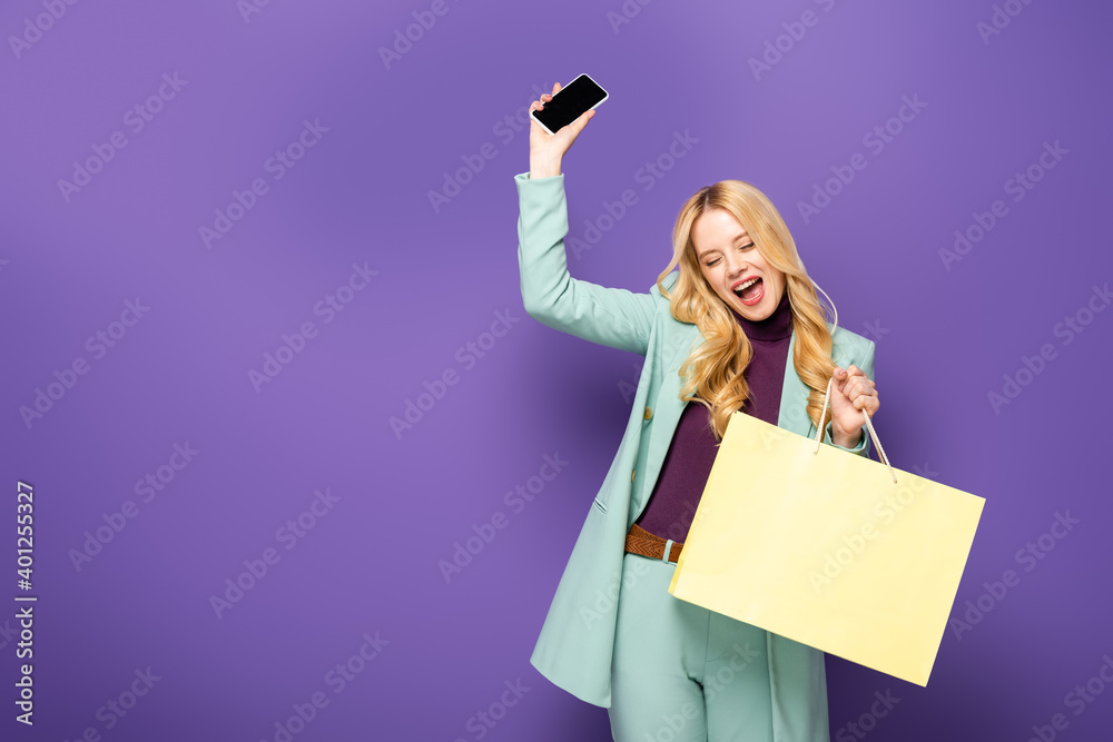 happy blonde young woman in fashionable turquoise blazer with smartphone and shopping bag on purple background. happy blonde young woman in fashionable turquoise blazer with smartphone and shopping bag on purple background.