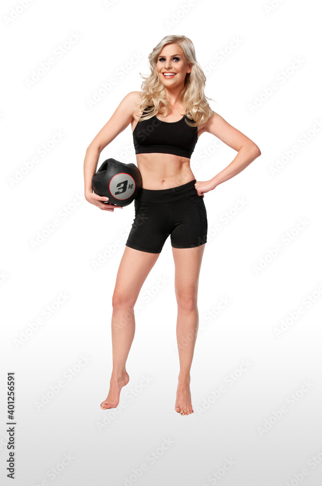 Portrait of a smiling young white female athlete with curly long blond hair posing by herself holding a black medicine ball in a studio with white background wearing black shorts & sports bra. Portrait of a smiling young white female athlete with curly long blond hair posing by herself holding a black medicine ball in a studio with white background wearing black shorts & sports bra.