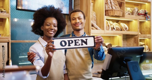 Portrait of cheerful young African American family married couple of bakers in aprons reopening bakehouse holding Open sign in hands. Small own bakery shop. Business concept photo