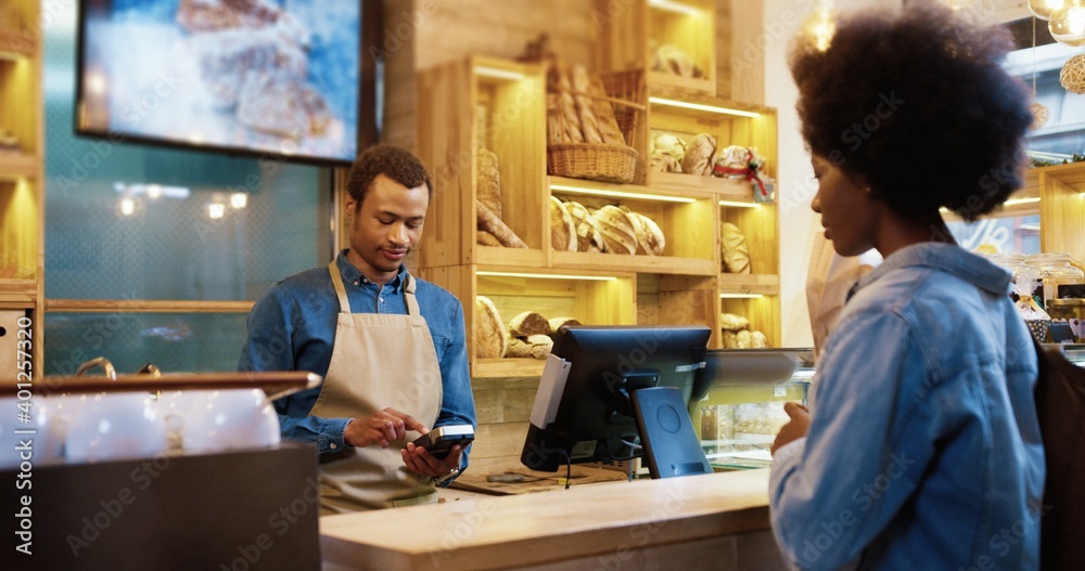 Close up of African American male seller in face mask and protective black gloves selling baked fresh bread in bakery shop. Client paying with credit card on device buying baking. Business concept