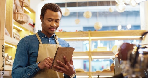 Close up of African American happy young handsome man worker in apron stands in bakery shop, texting and typing on tablet. Male seller browsing online on device. Business concept. Retail industry photo