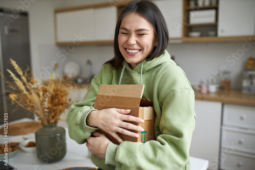 Cheerful teenage girl expressing excitement being overjoyed after receiving long awaited parcel with new clothing or accessories, embracing cardboard box, smiling broadly with her eyes closed photo