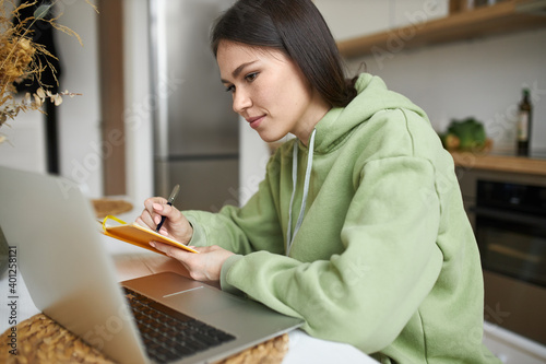 Stylish young dark haired woman in green hoodie sitting at table in front of open laptop, watching online investment course, learning how to manage finances, handwriting, putting down useful tips photo