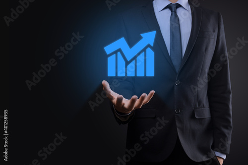 Businessman holding tablet and showing a growing virtual hologram of statistics, graph and chart with arrow up on dark background. Stock market. Business growth, planing and strategy concept.