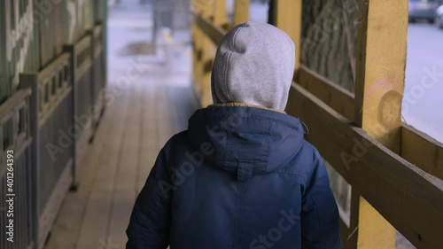 A homeless child waif walks along a fence in a slum. The view from the back. An orphan boy walks down the street photo