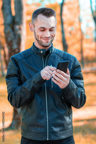 Fototapeta Naklejka Na Ścianę i Meble -  Youthful Satisfied Guy Using Black Smartphone at the Beautiful Autumn Park. Handsome Smiling Young Man with Mobile Phone at Sunny Day - Medium Shot Portrait