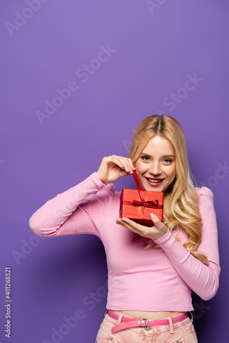 happy blonde young woman opening red gift box on purple background.