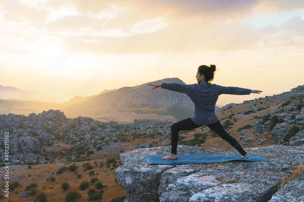 Full body of barefoot female standing in warrior pose on top of mountain while practicing yoga in nature at sunset time