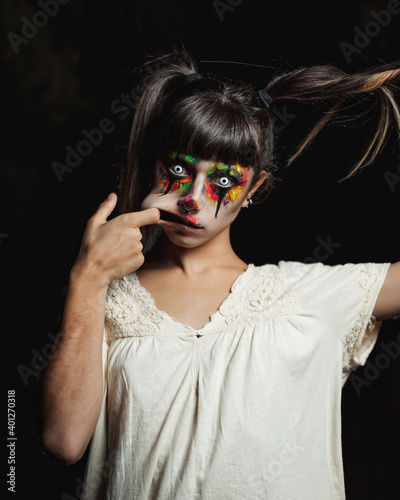 Scary young female in colorful contact lenses and with painted face standing in dark forest and smiling creepy while looking at camera on Halloween photo