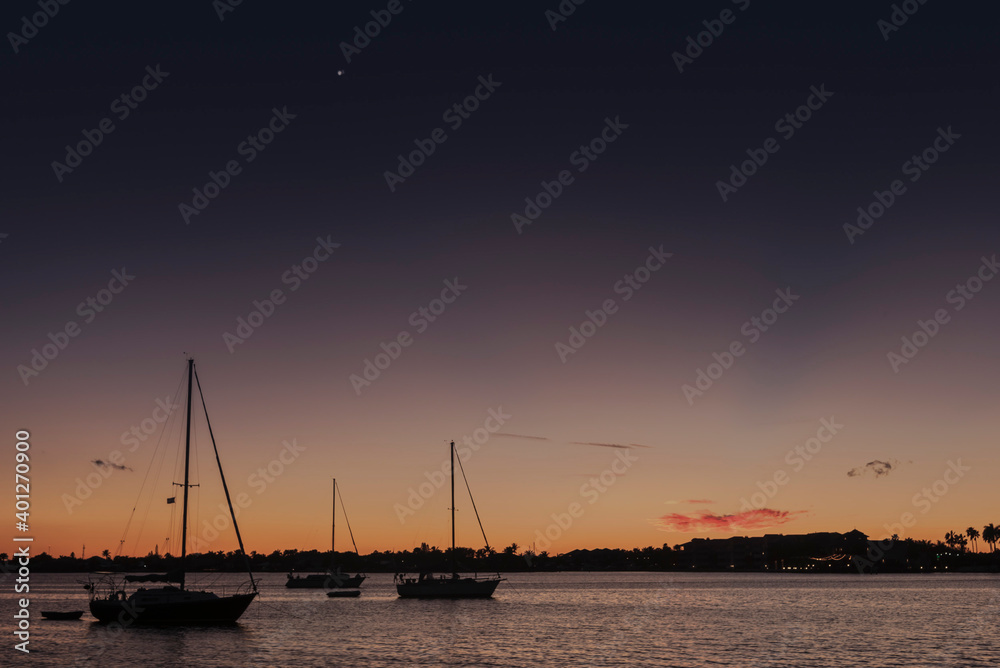 Moored sailboats silhouetted against a subtropical sunset with the planets Jupiter and Saturn aligned in the sky above