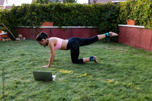 Side view of slim female athlete leaning on hand and knees while doing leg lifts and pumping hip muscles during workout wit h tutorial on the laptop with ankle weights in backyard in summer