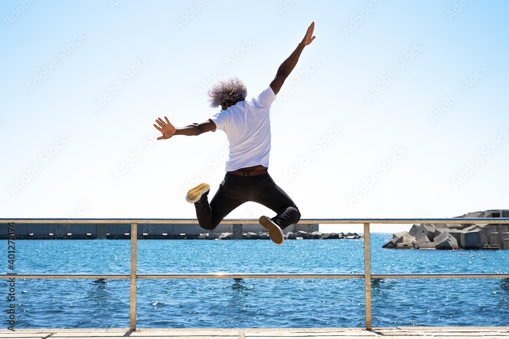 Black man with afro hair jumping in front of the sea and the beach. Concept of jumping in front of the sea. Blue tones.