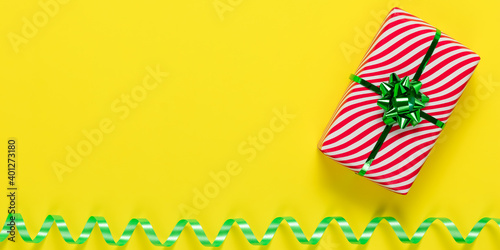 Yellow card and red striped white gift box with green bow and serpentine ribbon. Abstract wide banner with birthday present. Festive mockup design, holiday template with copy space. Panoramic frame.