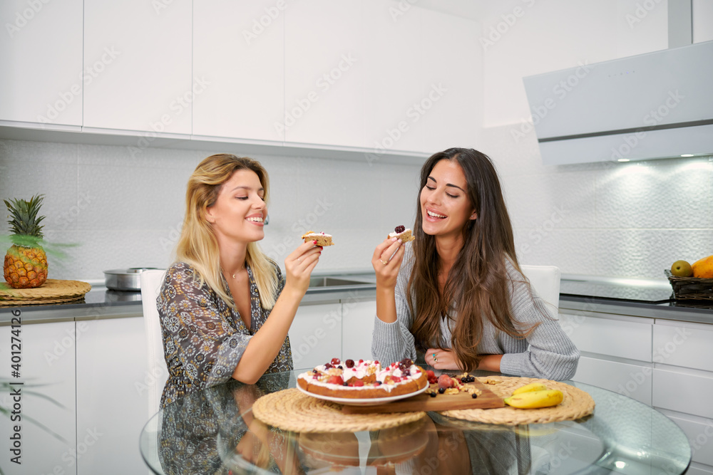 Cheerful young girlfriends sitting at table and enjoying delicious homemade vegan banana cake with cream and berries in home kitchen