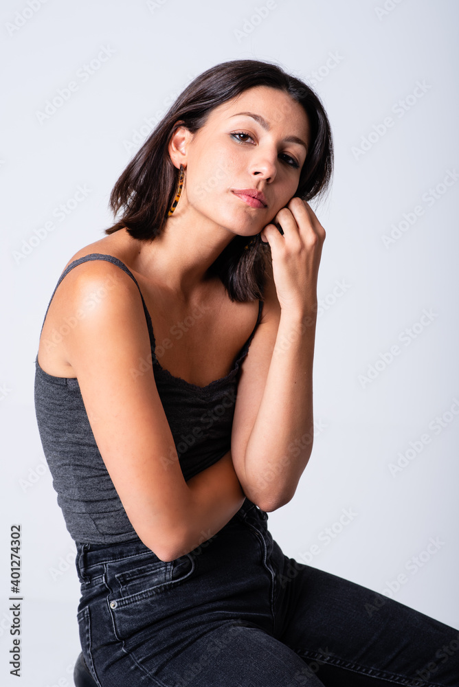 Full length young slim brunette in casual outfit sitting on stool against white background and looking at camera Full length young slim brunette in casual outfit sitting on stool against white background and looking at camera