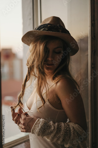 Young seductive female in stylish apparel and straw hat standing with closed eyes near window in apartment photo