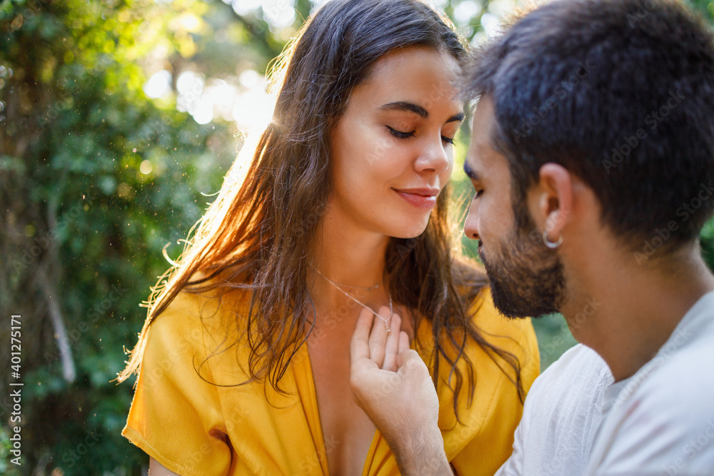 Side view of young bearded ethnic guy touching necklace of happy girlfriend while relaxing together in green garden on sunny day Side view of young bearded ethnic guy touching necklace of happy girlfriend while relaxing together in green garden on sunny day