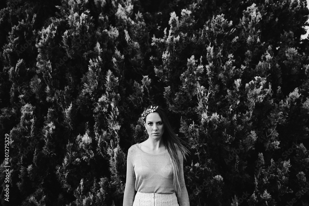 Confident young female wearing trendy hair wreath standing against abundant greenery while pointing finger gun and looking at camera Confident young female wearing trendy hair wreath standing against abundant greenery while pointing finger gun and looking at camera