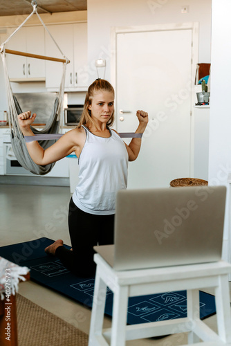 Sportswoman performing arms exercise with resistance bands while watching tutorials on laptop and working out at home
