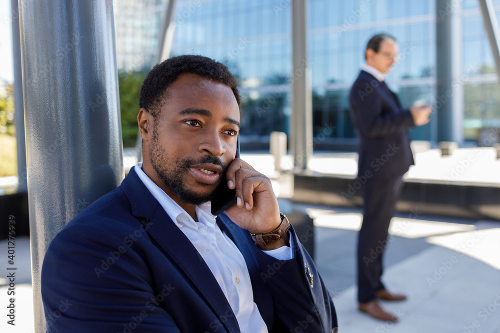 Confident young bearded African American  businessman in formal suit having phone conversation on street near contemporary building in sunlight