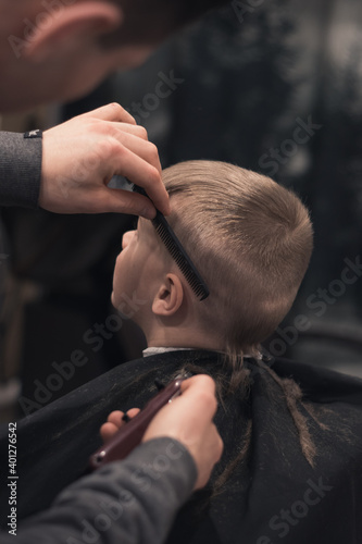 A pretty boy toddler happy to be on the haircut with a professional children's hairdresser. Blond little boy having a haircut at hair salon. Hairdresser's hands making hairstyle to child. © Iryna