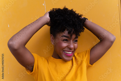 Happy young African American female with curly hair wearing bright yellow shirt and jeans standing against yellow wall and looking away