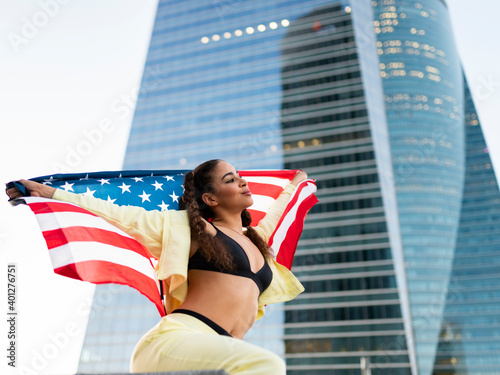 Side view of confident young ethnic lady in sports bra waving American flag and looking away against modern glass skyscraper photo