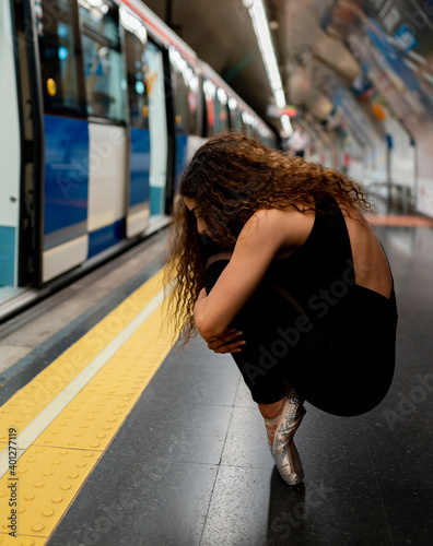 Side view of unrecognizable young ballerina with long curly hair in pointe shoes embracing knees while sitting on haunches in subway station photo