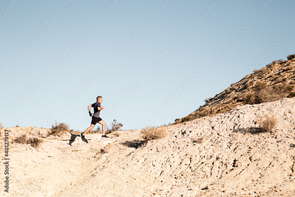 Side view of active male jogger running uphill in sandy semi desert terrain during workout in sunny morning Side view of active male jogger running uphill in sandy semi desert terrain during workout in sunny morning
