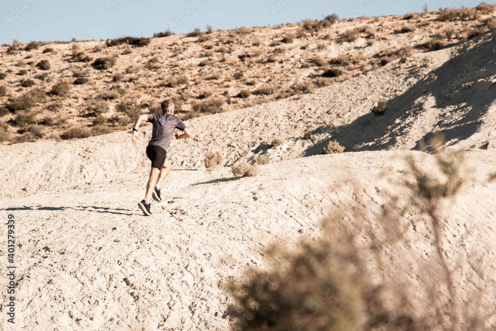 Back view of unrecognizable active male jogger running uphill in sandy semi desert terrain during workout in sunny morning Back view of unrecognizable active male jogger running uphill in sandy semi desert terrain during workout in sunny morning