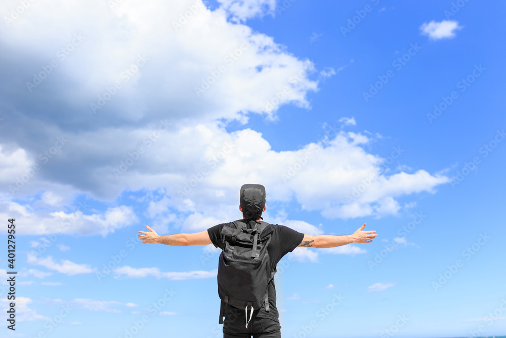 Low angle back view of male tourist in cap with backpack standing with raised arms under blue cloudy sky Low angle back view of male tourist in cap with backpack standing with raised arms under blue cloudy sky