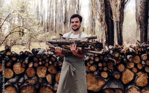 Man in apron taking logs from pile of firewood in forest for picnic