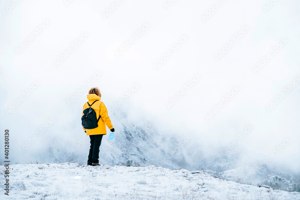 Back view of anonymous active traveler in bright yellow warm jacket with backpack holding trekking sticks and medical mask while standing on snowy hill admiring landscape Back view of anonymous active traveler in bright yellow warm jacket with backpack holding trekking sticks and medical mask while standing on snowy hill admiring landscape