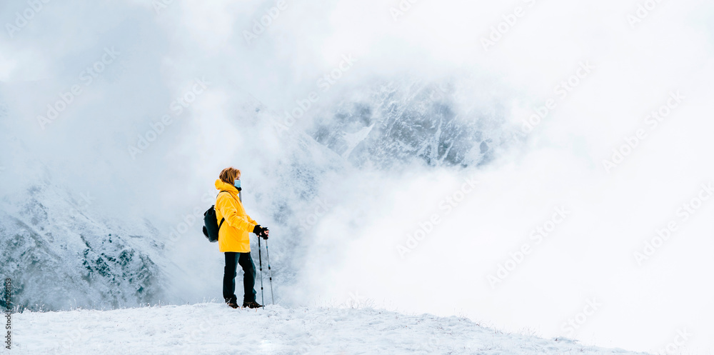 Side view of active traveler in bright yellow warm jacket with backpack holding trekking sticks and medical mask while standing on snowy hill admiring landscape Side view of active traveler in bright yellow warm jacket with backpack holding trekking sticks and medical mask while standing on snowy hill admiring landscape