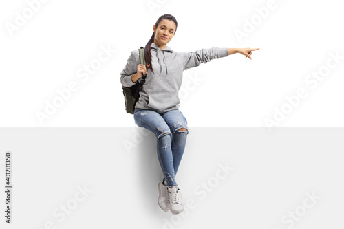 Cute female student sitting on a blank board and pointing to the side
