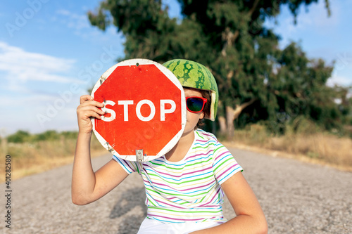 Stylish kid in ornamental watermelon head wear and sunglasses sitting on skateboard while showing signboard with STOP title on road photo