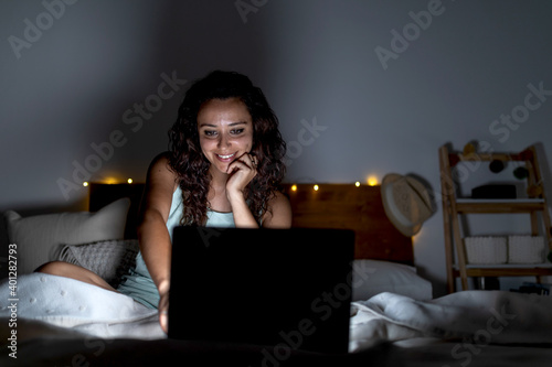 Delighted female in nightwear lying on bed in dark room and watching funny video on netbook while smiling and spending weekend at home photo