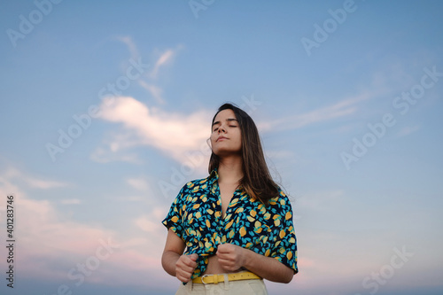 Low angle of young stylish mindful female with closed eyes under sky at sundown